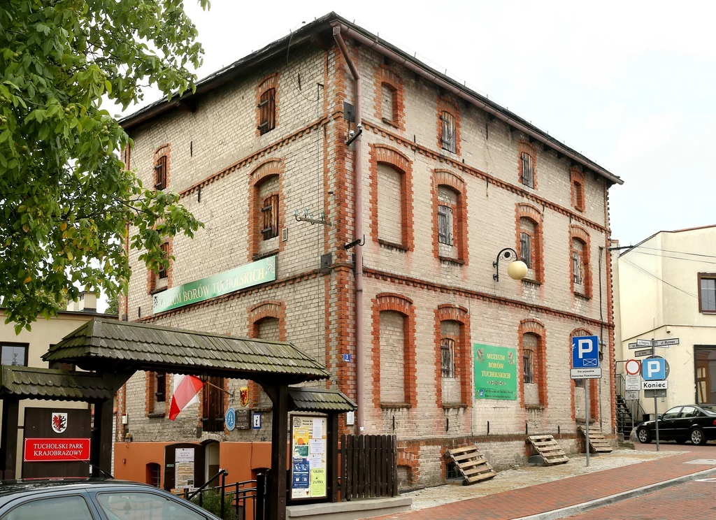 Muzeum Borów Tucholskich w Tucholi Ceglana kamienica o trzech kondygnacjach z zamurowanymi oknami, na której wiszą dwa zielone banery reklamowe, obok znajduje się budka informacyjna z daszkiem oraz widoczne znaki parkingowe.