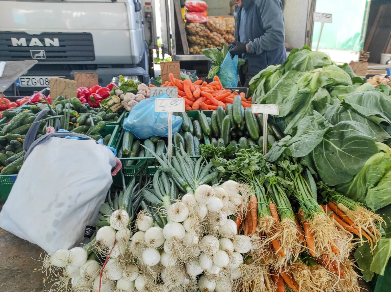 Stoisko z warzywami na targu z wyeksponowanymi marchewkami, ogórkami, białą cebulą, sałatą i innymi świeżymi produktami, za nimi widoczna ciężarówka i sprzedawca.