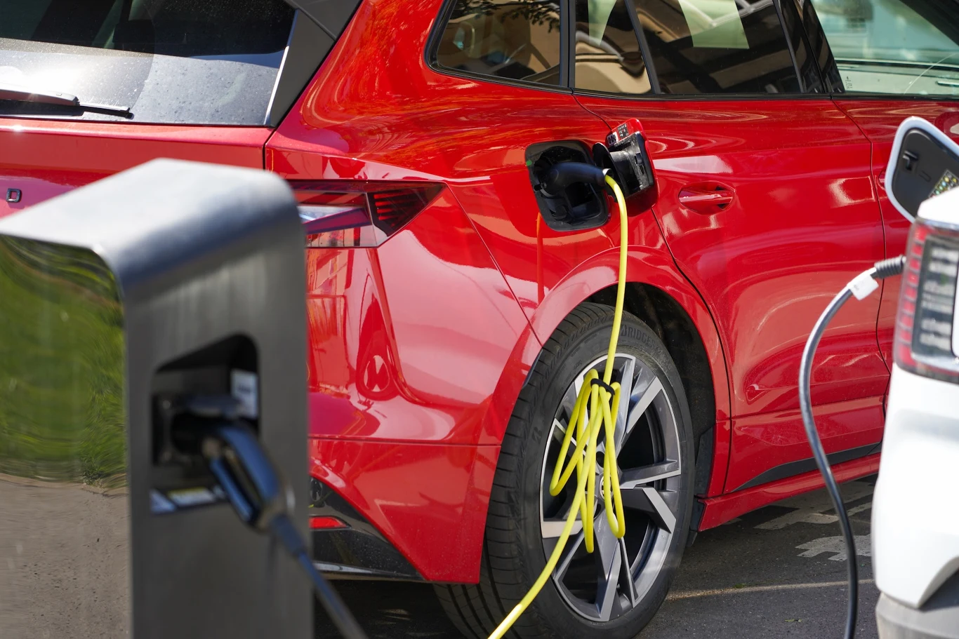 Polacy kupują samochody elektryczne, ale wciąż nie dostali ani złotówki dofinansowania Czerwony samochód elektryczny podłączony do stacji ładowania, widoczna żółta ładowarka, fragment sąsiedniego pojazdu oraz element infrastruktury do ładowania aut elektrycznych.