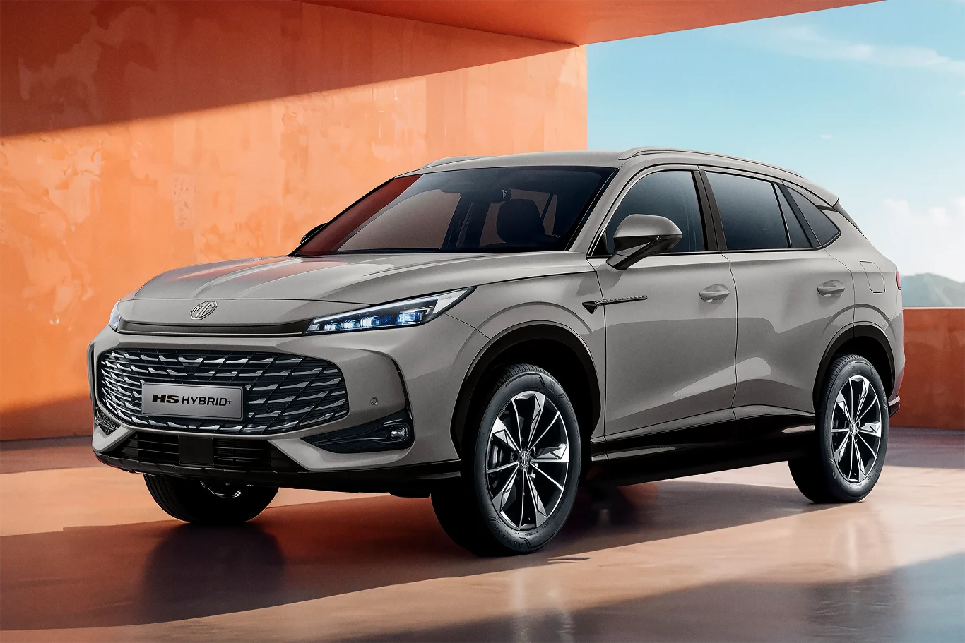 MG HS Hybrid+ debiutuje na polskim rynku Nowoczesny samochód SUV typu hybrid w odcieniu szarości, stojący na tle pomarańczowej ściany w dobrze oświetlonym miejscu. Duże felgi, charakterystyczny grill oraz logo producenta podkreślają nowoczesny charakter pojazdu.