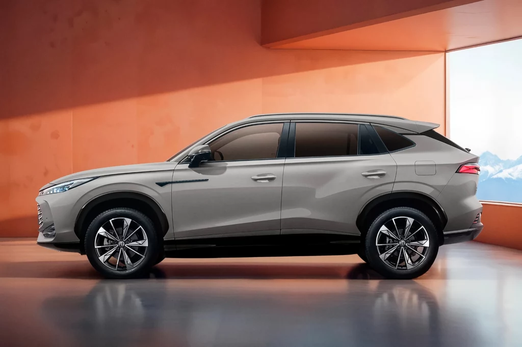 MG HS Hybrid+ ma 224 KM i spala w trasie 5,5 l/100 km Nowoczesny samochód SUV w kolorze szarym w profilu bocznym, osadzony w minimalistycznym, pomarańczowym wnętrzu z dużym oknem, przez które widać fragment górskiego krajobrazu.