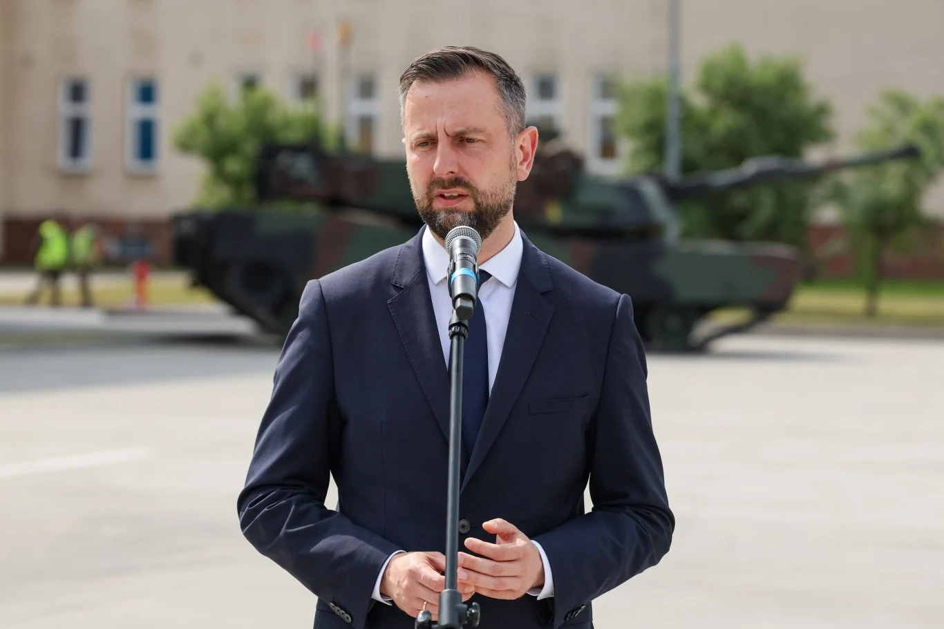 Władysław Kosiniak-Kamysz wziął udział w uroczystościach ku pamięci sierż. Mateusza Sitka Mężczyzna w garniturze wygłasza przemówienie przy mikrofonie na tle nowoczesnych czołgów ustawionych na placu o jasnej nawierzchni, w tle budynek i drzewa.