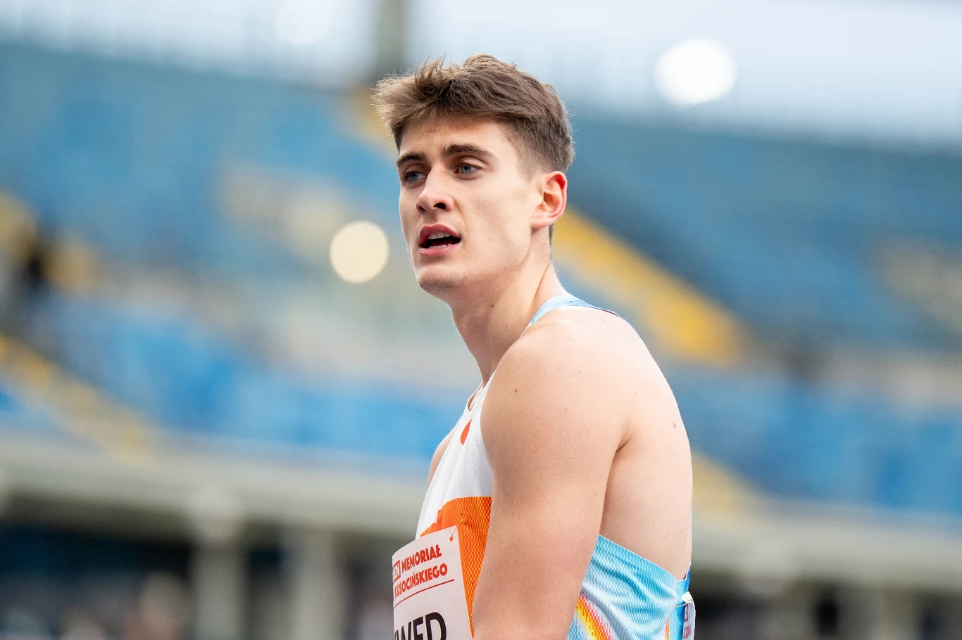 Młody sportowiec w lekkoatletycznym stroju na stadionie, patrzący w dal, z wyrazem skupienia na twarzy, w tle rozmyte niebieskie trybuny.