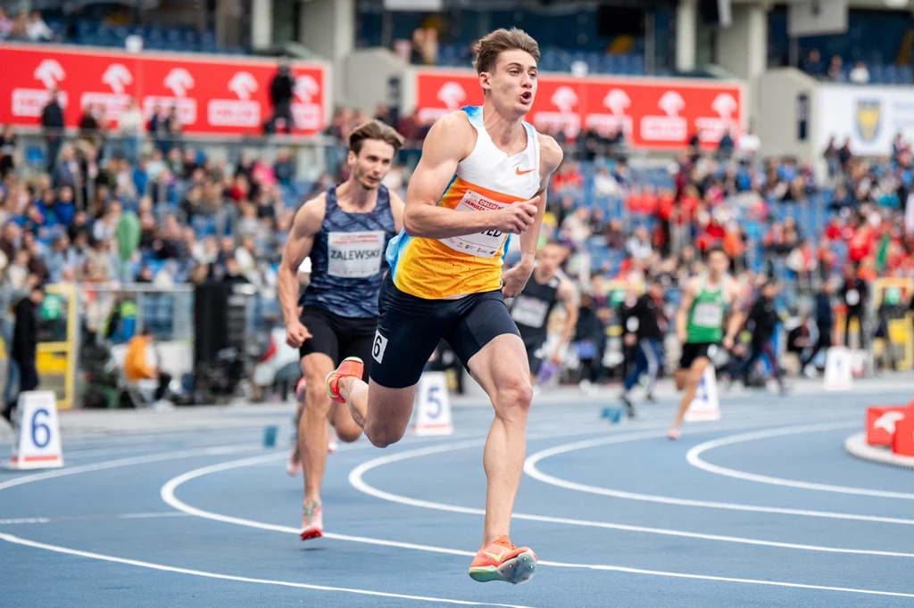 Maksymilian Szwed Zawodnicy rywalizują na bieżni podczas zawodów lekkoatletycznych, na pierwszym planie sportowiec w żółto-białej koszulce biegnie jako lider, w tle kibice na trybunach oraz inni uczestnicy biegu.