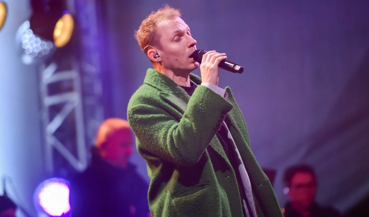 Igor Herbut był jedną z gwiazd tegorocznych "Wianków nad Wisłą" Mężczyzna w zielonym płaszczu śpiewa do mikrofonu na scenie, w tle oświetlenie sceniczne i zarys innych osób.
