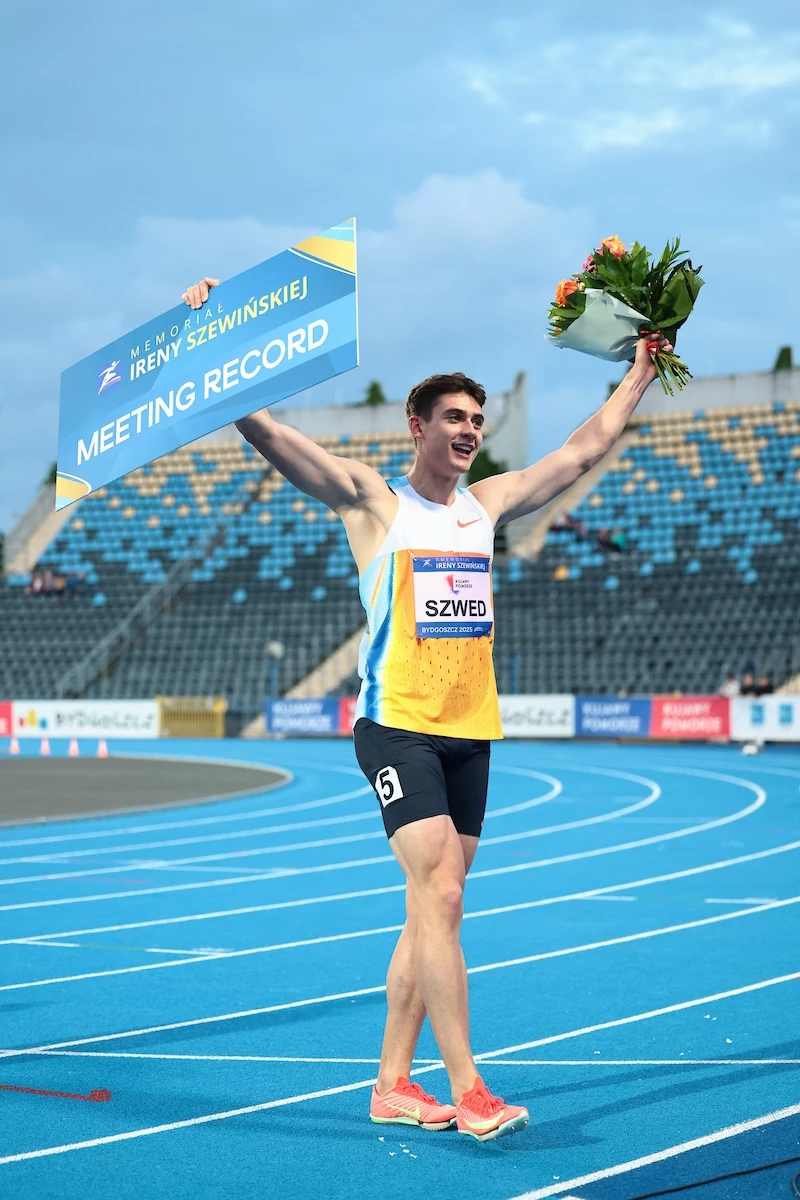 Maksymilian Szwed Sportowiec w stroju lekkoatletycznym triumfalnie unosi tablicę z napisem 'Meeting Record' oraz bukiet kwiatów na stadionie lekkoatletycznym, na trybunach widoczni kibice.