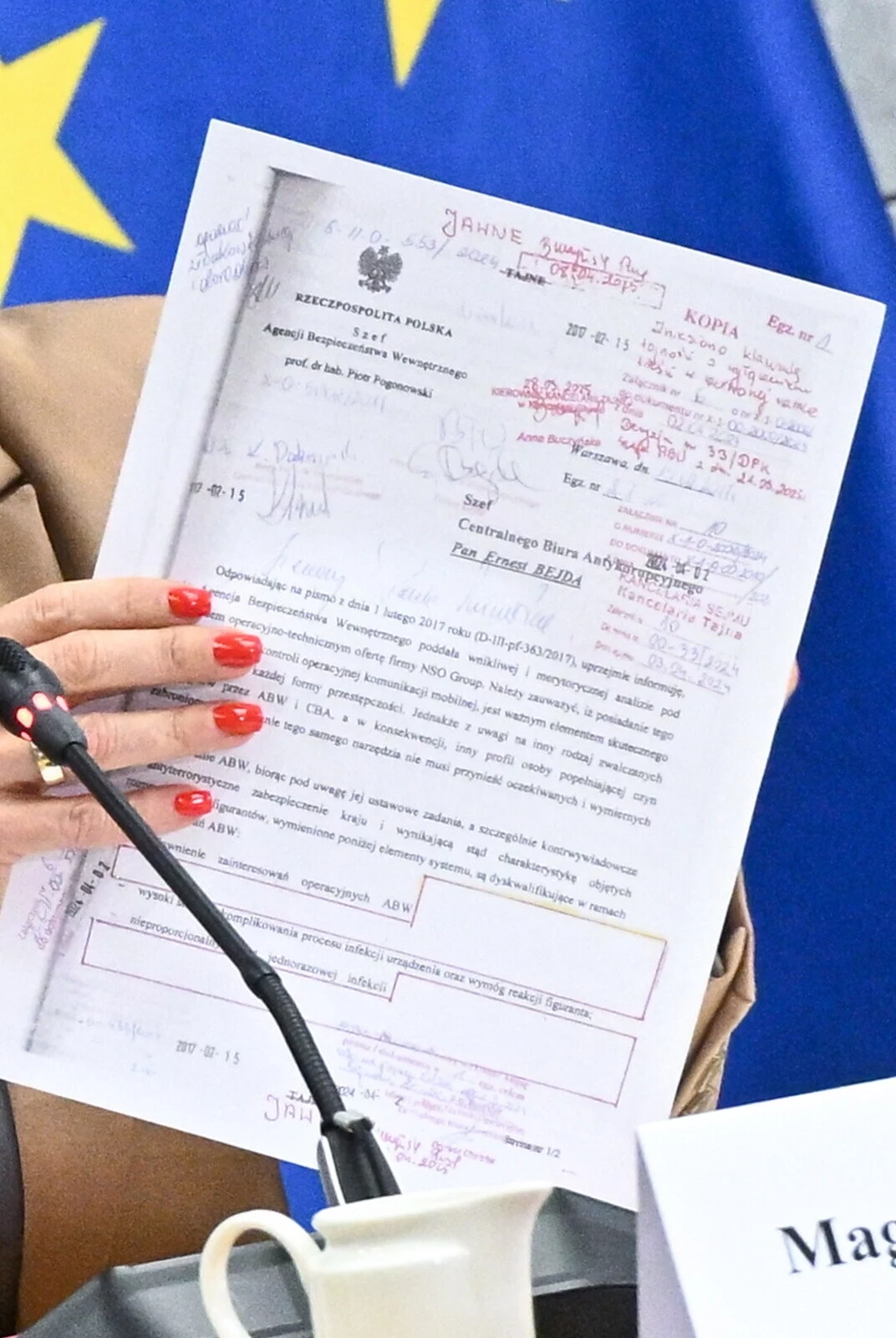 Odtajniony dokument na komisji śledczej ds. Pegasusa. Pokazano opinię ABW wysłaną do ówczesnego szefa CBA Kobieta o czerwonych paznokciach trzyma polski urzędowy dokument przed mikrofonem, na tle niebieskiej flagi z żółtymi gwiazdami, na stole znajduje się filiżanka i tabliczka z napisem.