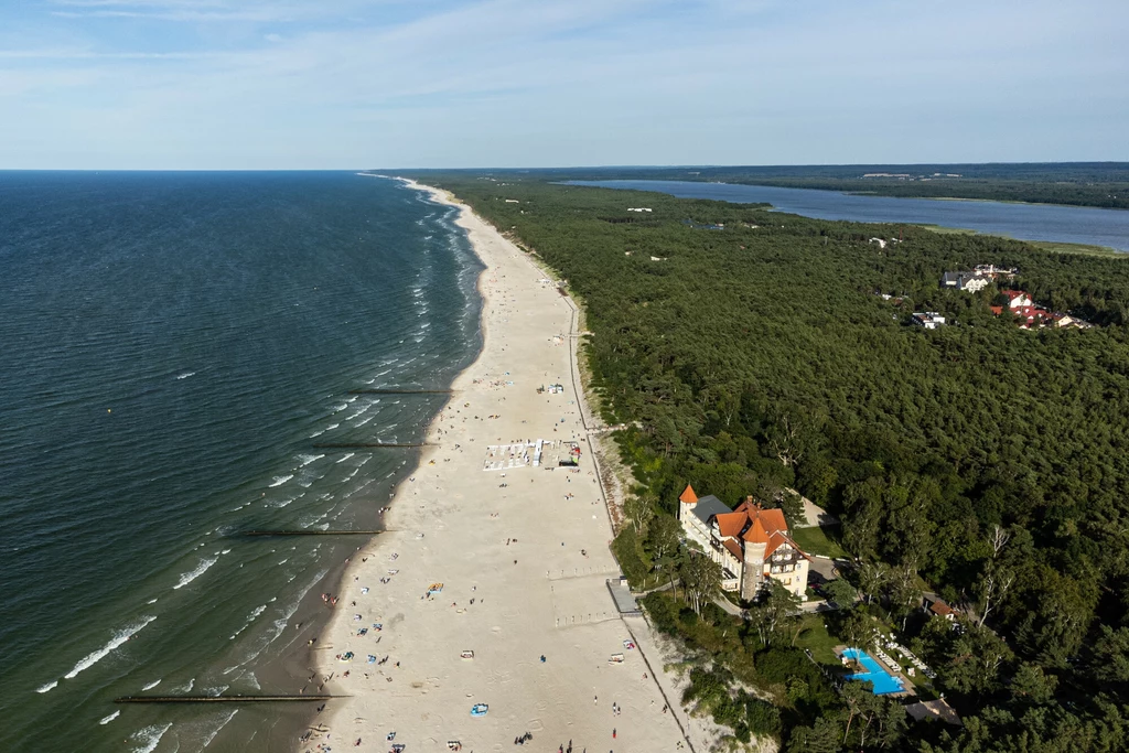 Położona między dwoma jeziorami Łeba dostarczy mnóstwa atrakcji. Nie będziesz się nudzić nawet w niepogodę Piękna, szeroka plaża ciągnąca się wzdłuż linii brzegowej Bałtyku, obok gęsty las i elegancki budynek z czerwonym dachem, wokół którego rozpościera się zielony ogród i niewielki basen, w tle widać rozległe tereny leśne oraz spokojne wody zatoki.