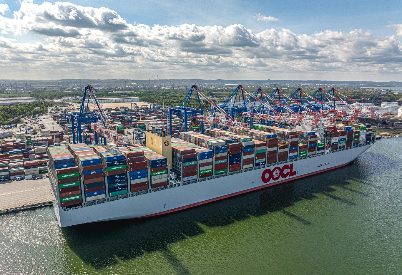 Baltic Hub przyjmie największe statki kontenerowe świata Ogromny kontenerowiec załadowany kolorowymi kontenerami cumuje przy nabrzeżu portowym, w tle widoczne dźwigi portowe i rozległa infrastruktura portowa pod błękitnym niebem.