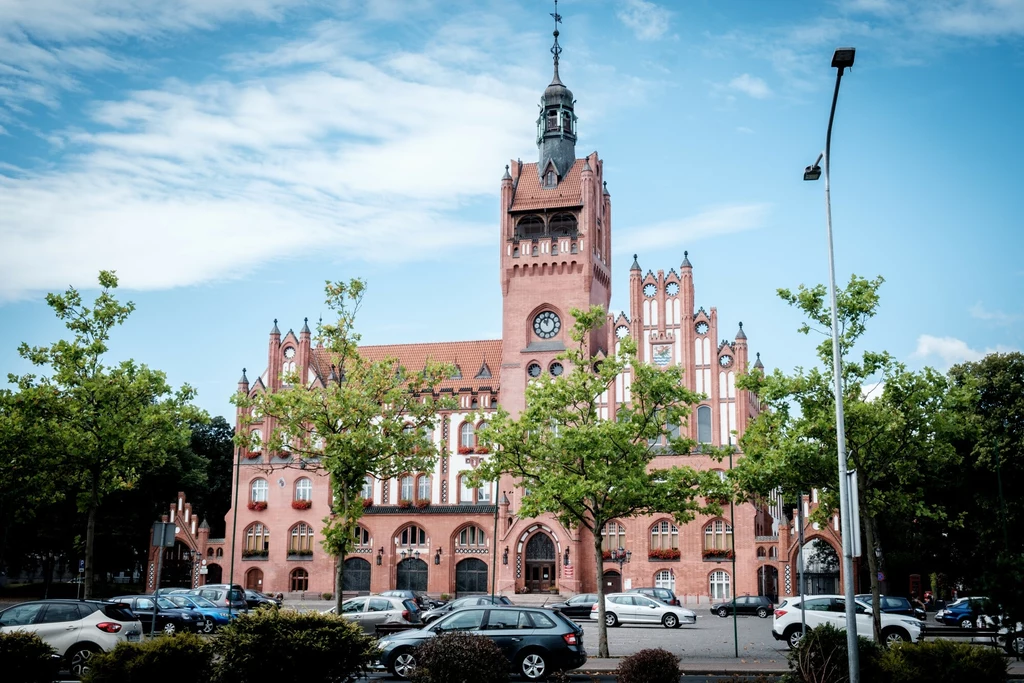 Ratusz w Słupsku to jeden z najpiękniejszych zabytków. Miasto oferuje jednak znacznie więcej atrakcji i jest świetną bazą wypadową do doliny Słupi Ceglany budynek ratusza w stylu neogotyckim z wysoką wieżą zegarową, otoczony zielonymi drzewami, przed którym znajduje się parking z zaparkowanymi samochodami i nowoczesnymi latarniami ulicznymi.