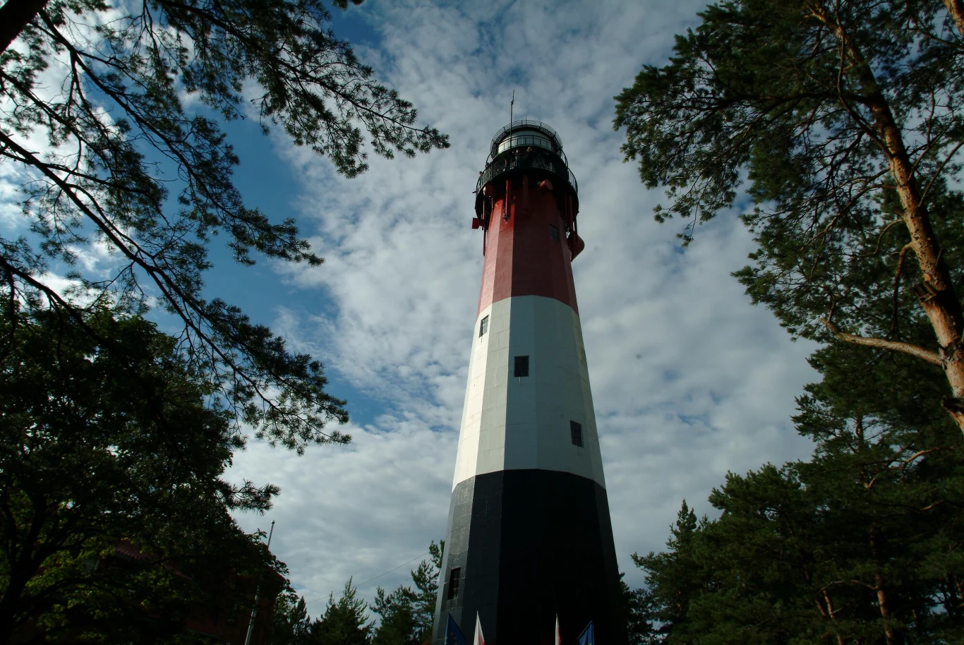 Wysoka latarnia morska o czerwono-biało-czarnej elewacji otoczona drzewami pod niebem częściowo pokrytym chmurami.
