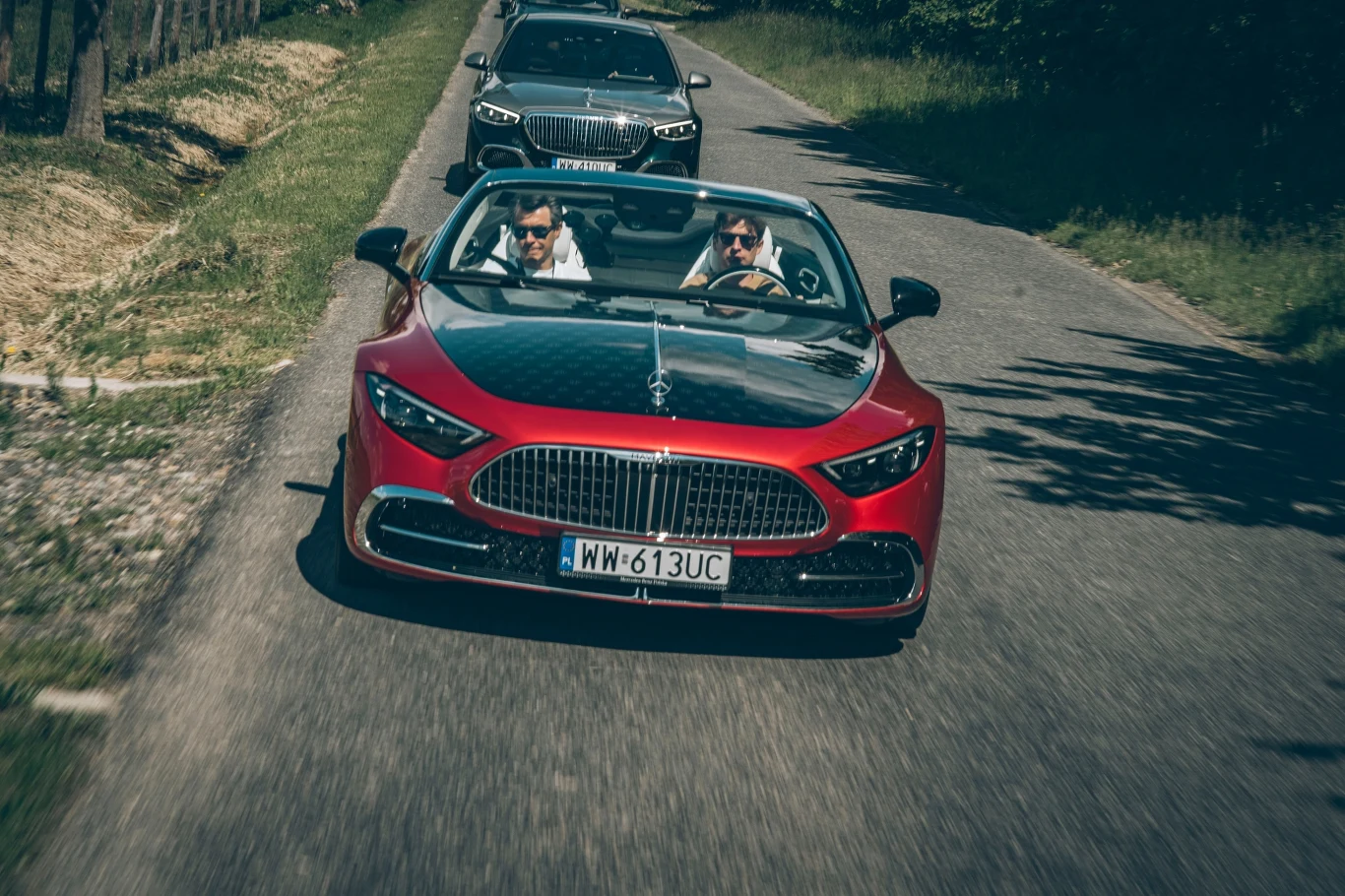 Mercedes-Maybach SL 680 Monogram Series to aktualnie najdroższy model marki w gamie. Lista opcji kończy się na wyborze obręczy i kolorów nadwozia Czerwony kabriolet z dwoma mężczyznami w środku jadący wiejską asfaltową drogą, w tle ciemny samochód osobowy, słoneczna pogoda, droga otoczona drzewami i ogrodzeniem po lewej stronie.