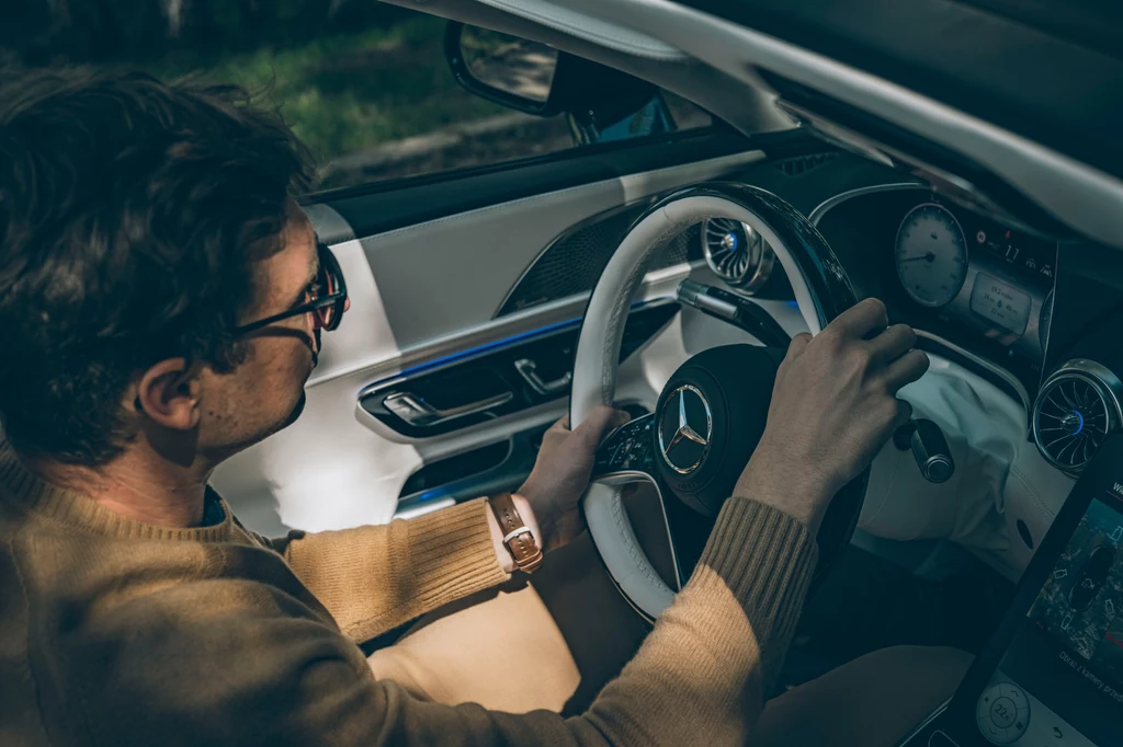 Ręcznie wyszywana kierownica powstaje przez jeden dzień. W wersji Maybach nie można wybrać innego wykończenia niż jasne z ciemnym, lakierowanym drewnem Mężczyzna prowadzący luksusowy samochód marki Mercedes, skupiony na jeździe, z rękami na kierownicy oraz widocznymi nowoczesnymi elementami wnętrza pojazdu.