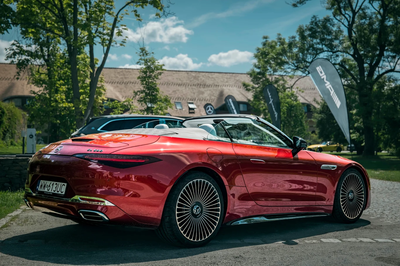 Opcjonalne, widoczne na zdjęciach obręcze mają rozmiar 21 cali i kosztują prawie 29 tys. zł ekstra Czerwony kabriolet marki Mercedes-AMG zaparkowany na tle zielonych drzew i budynku, błyszcząca karoseria odbija światło słoneczne, a w tle widoczne są banery eventowe i drugi samochód.