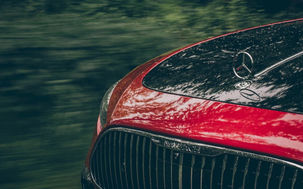SL 680 Maybach to jeden z nielicznych modeli marki z klasyczną, wystającą gwiazdą na masce Czerwona maska samochodu Mercedes z widocznym znakiem firmowym oraz grillem, na powierzchni widoczne odbicia otoczenia i krople deszczu.