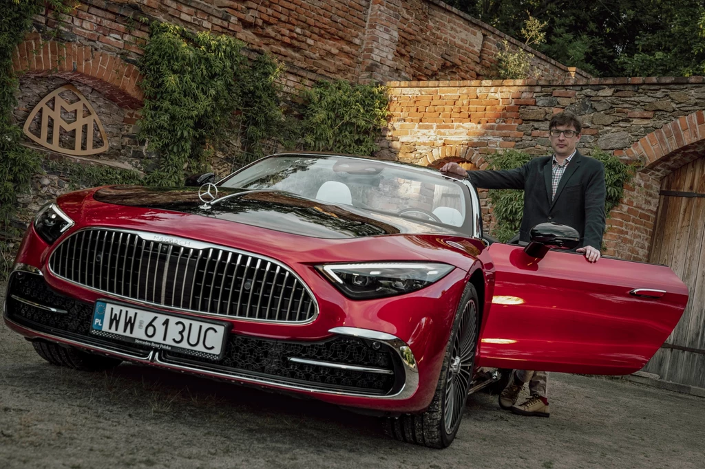 Nadwozie wyróżnia się dużą ilością chromu oraz licznymi oznaczeniami - są między innymi w zderzaku czy reflektorach Czerwony kabriolet marki Mercedes-Maybach zaparkowany na tle ceglanych murów, obok samochodu stoi elegancko ubrany mężczyzna z otwartymi drzwiami kierowcy, roślinność porastająca mur i wyeksponowane detale auta przyciągają uwagę.