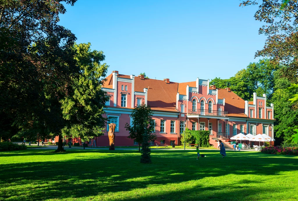 Klasycystyczny pałac w Wejherowie otoczony jest pięknym parkiem, przez który przepływa Cedron Różowy pałac o bogatej architekturze otoczony zielonym parkiem pełnym trawników, dużych drzew i krzewów, z białymi parasolami ogrodowymi i ludźmi spacerującymi na tle jasnego nieba.