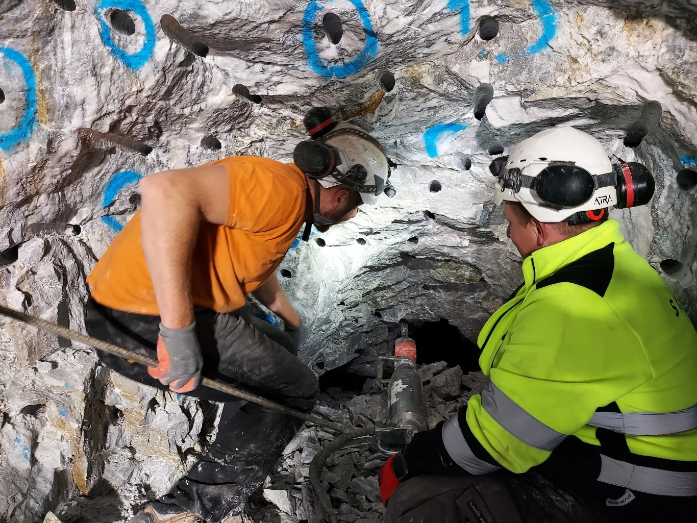 Przebili się do Sali Mastodonta w Jaskini Niedźwiedziej! Dwóch górników w ochronnych kaskach i słuchawkach prowadzi prace w tunelu wykutym w skale, otoczeni są narzędziami oraz skałami oznaczonymi niebieskim sprayem.