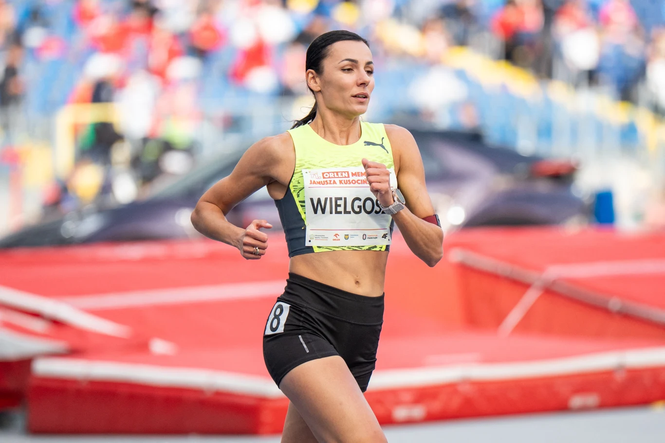 Atletka w żółtym stroju sportowym biegnie po bieżni stadionu, z numerem startowym przypiętym do koszulki, w tle widoczne trybuny i rozmazani kibice.