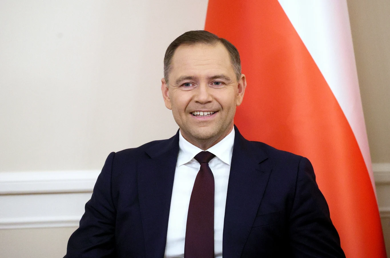 Prezydent Karol Nawrocki Uśmiechnięty mężczyzna w garniturze i bordowym krawacie siedzi na tle biało-czerwonej flagi i jasnej ściany.
