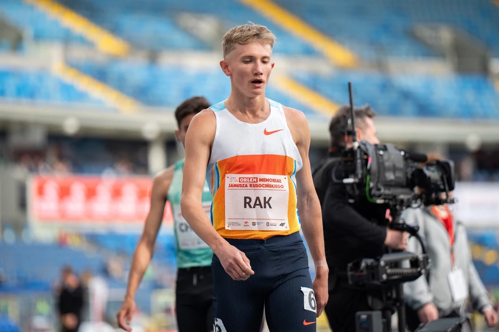 Filip Rak Lekkoatleta w stroju sportowym z numerem startowym i nazwiskiem uczestnika stoi na bieżni stadionu, z tyłu widoczni inni zawodnicy oraz ekipa z kamerą filmową, trybuny są częściowo puste.