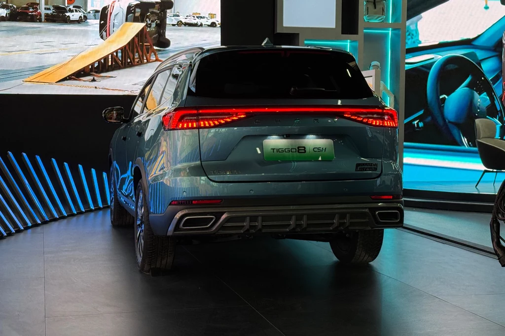 Tylne lampy Chery Tiggo 8 łączy świetlna listwa Tył niebieskiego SUV-a Chery Tiggo 8 stoi na ekspozycji targowej, otoczony podświetlonymi elementami stoiska, na ekranie w tle widoczny fragment toru testowego z rampą.