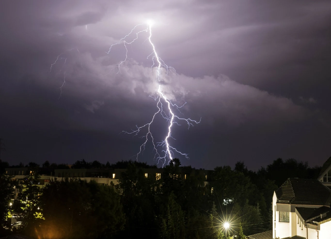 Silne uderzenie błyskawicy rozświetlające nocne niebo nad zabudowaniami, w tle widoczne ciemne chmury oraz korony drzew oświetlone słabym światłem ulicznych lamp.