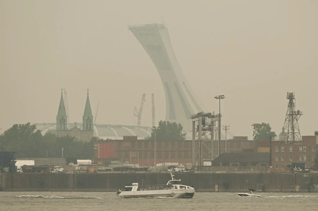 Zamglona panorama miasta ze stadionem i charakterystyczną wieżą w tle, na pierwszym planie widoczne są łodzie płynące po rzece, zabudowa przemysłowa oraz słabe światło i ograniczona widoczność.