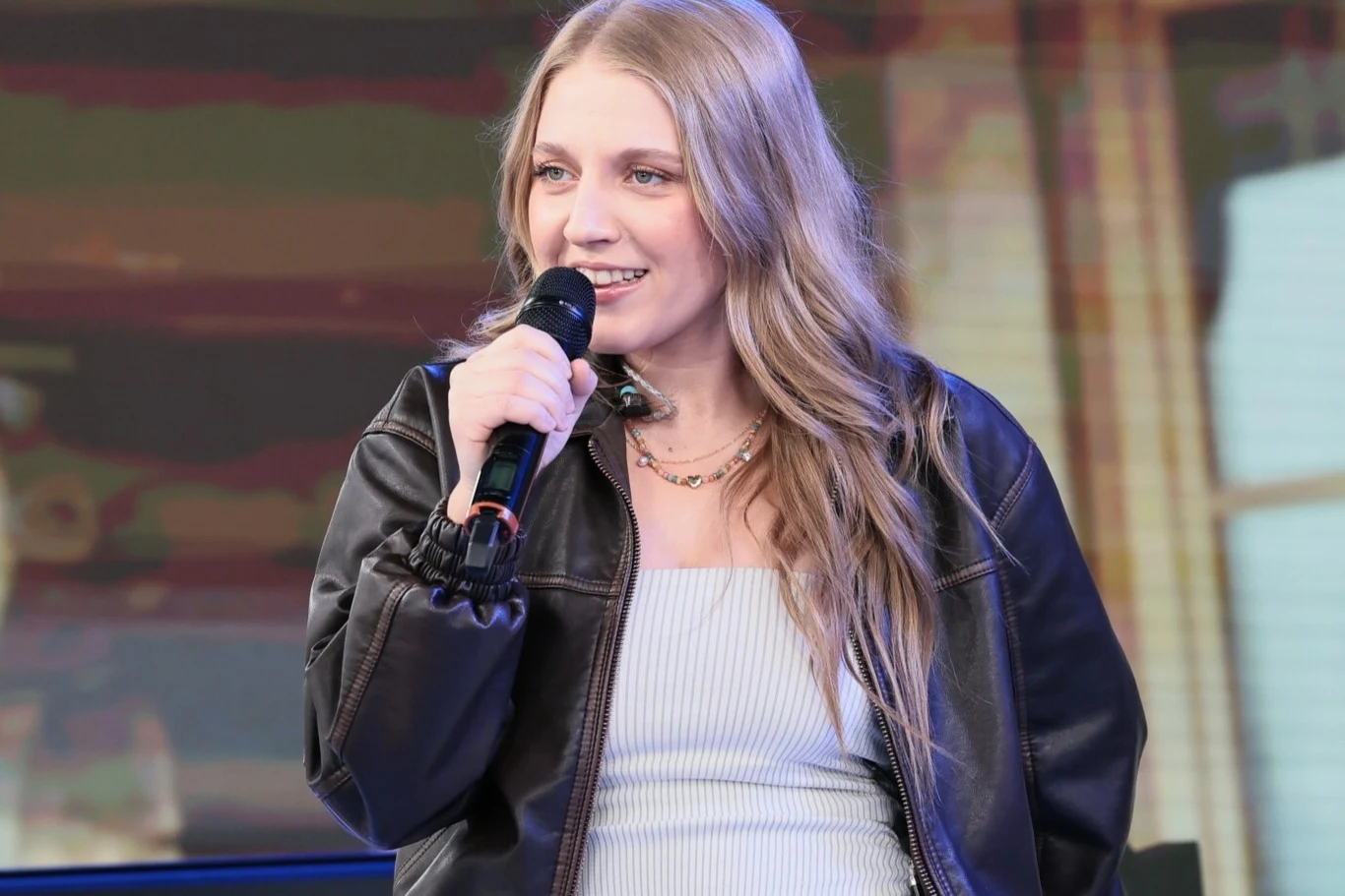 Maja Golec pójdzie w ślady znanego taty? Młoda kobieta z długimi blond włosami trzyma mikrofon i uśmiecha się podczas występu na scenie, ubrana w czarną skórzaną kurtkę i jasny top, w tle rozmyte światło i fragmenty dekoracji scenicznej.