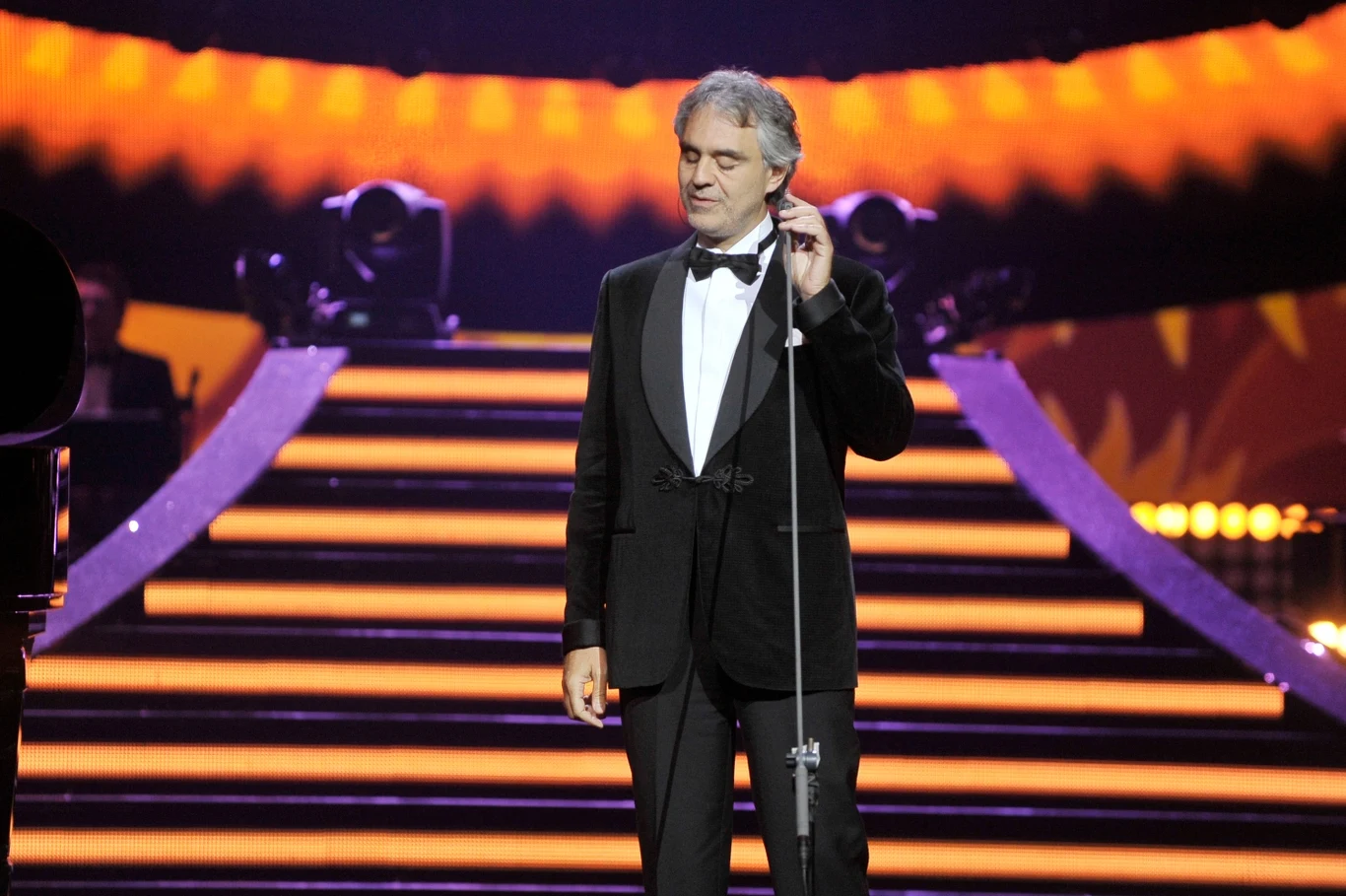 Andrea Bocelli zagra w Polsce Elegancko ubrany mężczyzna w smokingu śpiewający na scenie, w tle podświetlone schody z pomarańczowym oświetleniem i reflektorami, atmosfera koncertu lub gali