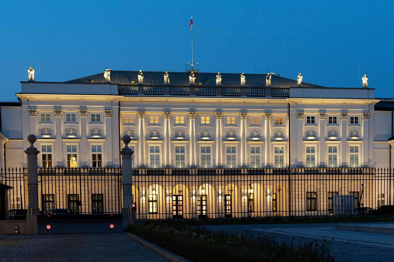 Pałac Prezydencki Oświetlona fasada pałacu z symetrycznie rozmieszczonymi oknami, rzędem kolumn, rzeźbami na dachu oraz flagą na szczycie budynku, okolona ogrodzeniem, wieczorna sceneria podkreśla elegancję architektury.