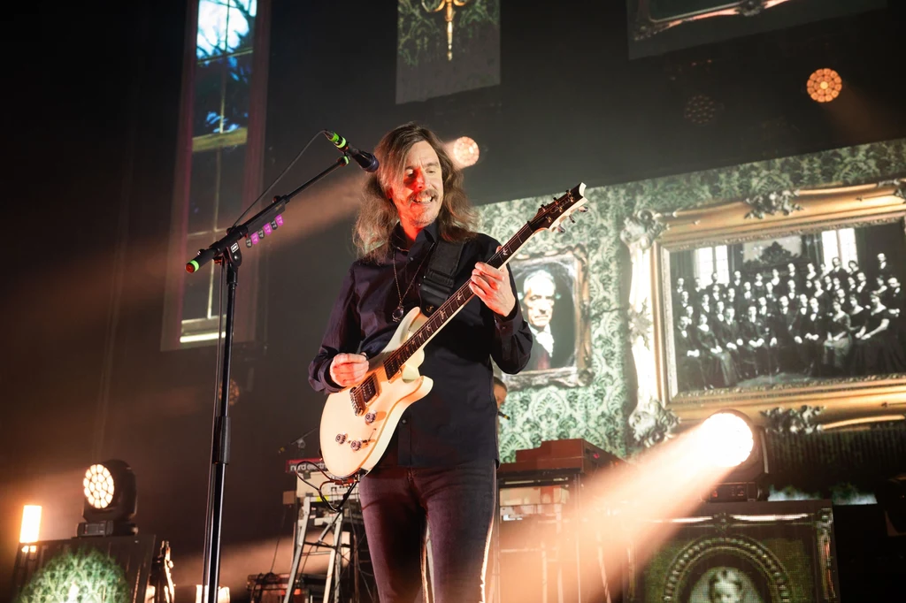 Opeth Muzyk z długimi włosami grający na białej gitarze elektrycznej podczas koncertu, w tle podświetlone ramki z czarno-białymi zdjęciami i rozproszonym oświetleniem scenicznym.