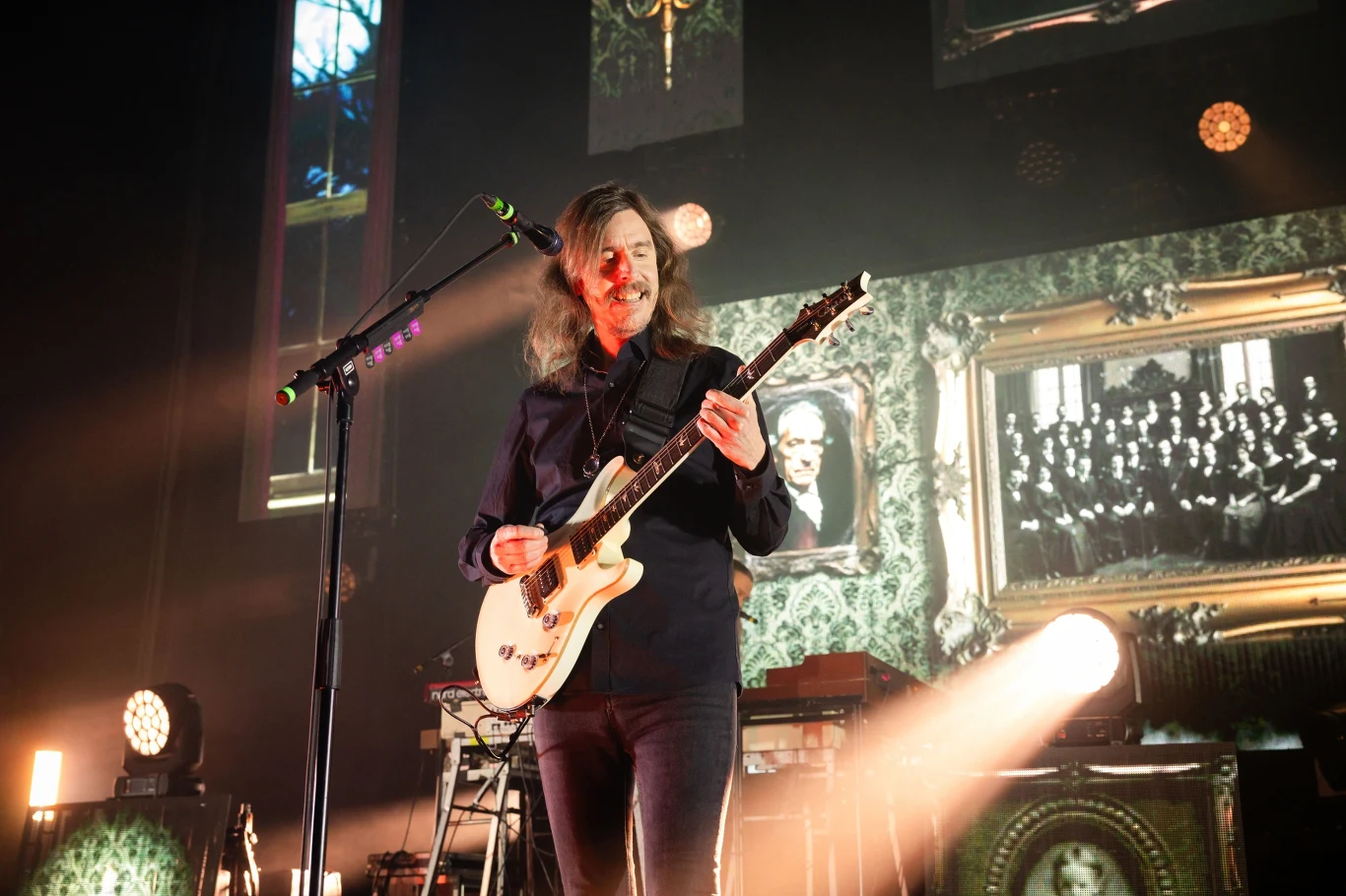 Muzyk z długimi włosami grający na białej gitarze elektrycznej podczas koncertu, w tle podświetlone ramki z czarno-białymi zdjęciami i rozproszonym oświetleniem scenicznym.