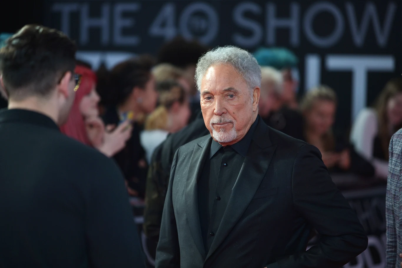Tom Jones ma dwóch synów Starszy mężczyzna w eleganckim czarnym garniturze stoi na tle rozmytego tłumu podczas wydarzenia oświetlonego scenicznie, patrząc spokojnie przed siebie.
