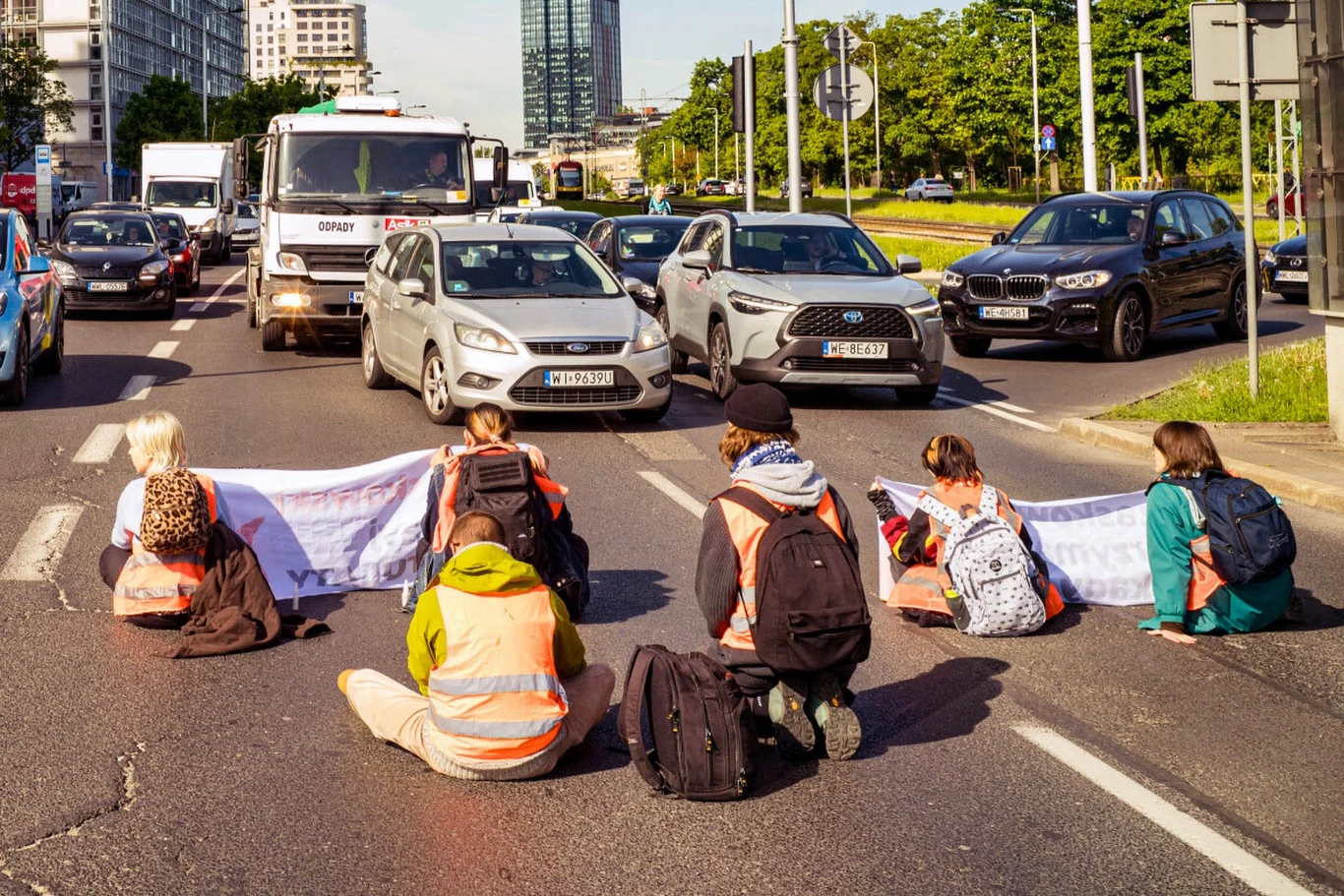 Od teraz za podobny protest we Włoszech będzie można trafić do więzienia na 2 lata Grupa osób siedzących na środku ulicy z rozwiniętym transparentem, blokująca ruch samochodów na wielopasmowej ulicy w dużym mieście w otoczeniu nowoczesnych budynków i zieleni.