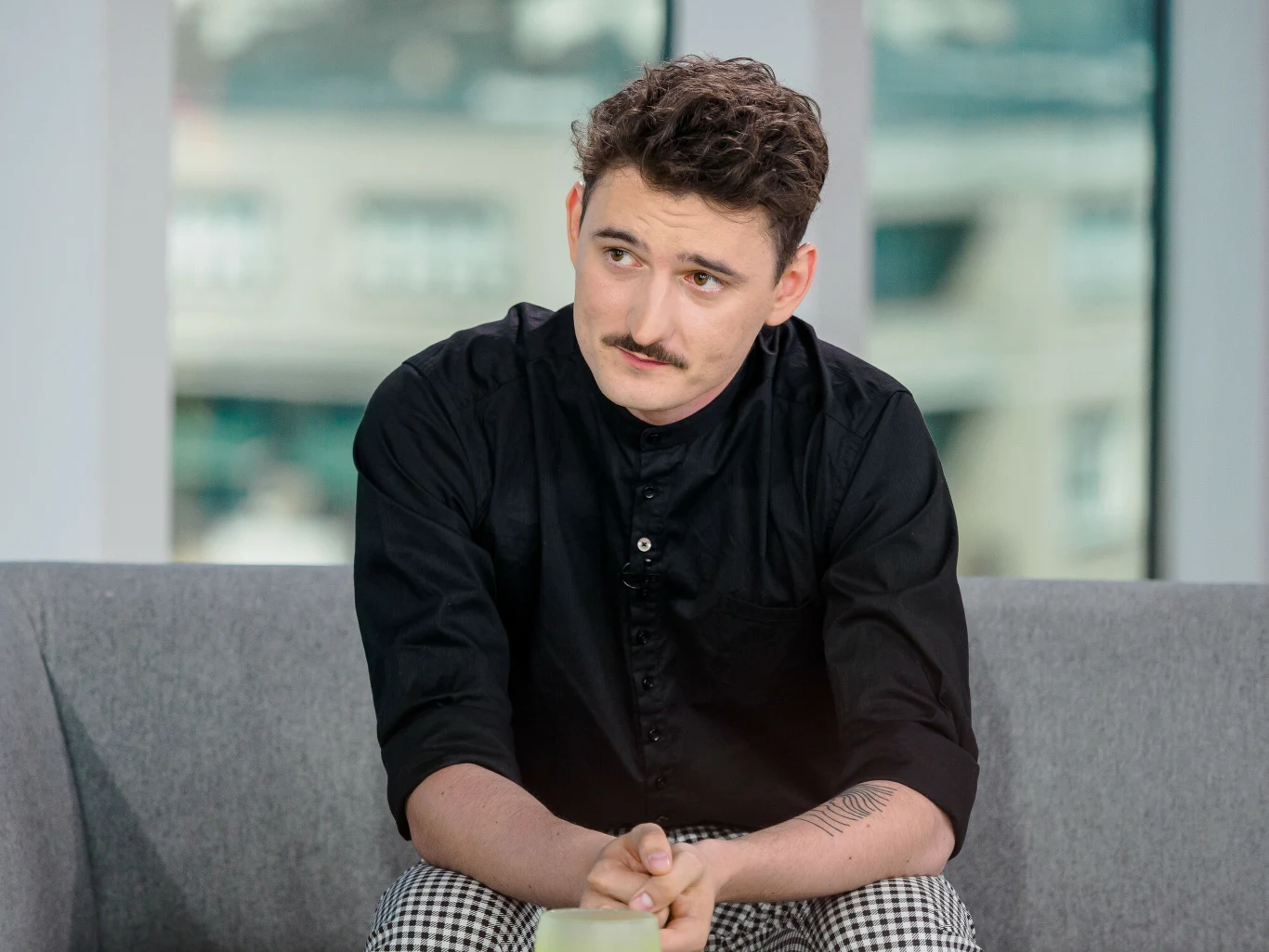 Dawid Podsiadło zorganizował Festiwal Zorza Młody mężczyzna z krótkimi, kręconymi włosami i wąsami siedzi na szarej sofie w jasnym pomieszczeniu, ubrany w czarną koszulę i kraciaste spodnie, patrzy lekko w bok trzymając ręce splecione.