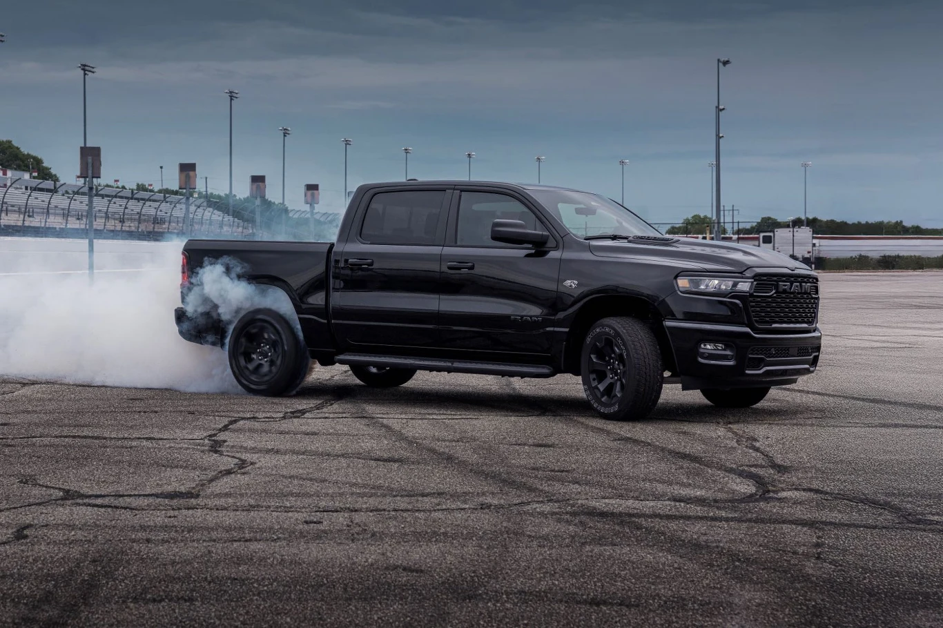 Czarny pickup marki RAM na torze, tylne koła generują dym podczas gwałtownego przyspieszenia lub driftu, asfalt z wyraźnymi śladami opon, w tle barierki toru oraz słupy oświetleniowe pod pochmurnym niebem.