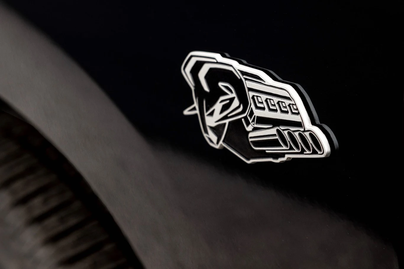 Metalowy emblemat przedstawiający stylizowany wizerunek silnika umieszczony na czarnej karoserii pojazdu, obok widoczna jest część opony.