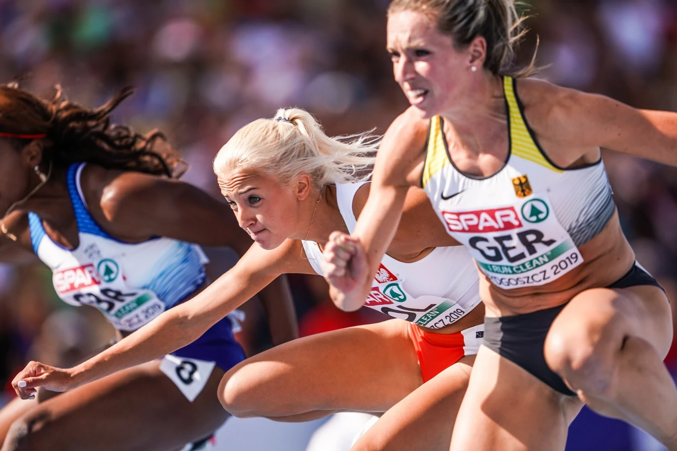 Karolina Kołeczek, siedmiokrotna mistrzyni Polski na 100 m przez płotki, ogłosiła zakończenie kariery Trzy sprinterki w sportowych strojach rywalizują w wyścigu na profesjonalnym stadionie lekkoatletycznym, wszystkie są skupione i gotowe do startu, tłum w tle podkreśla ogromne emocje sportowe.