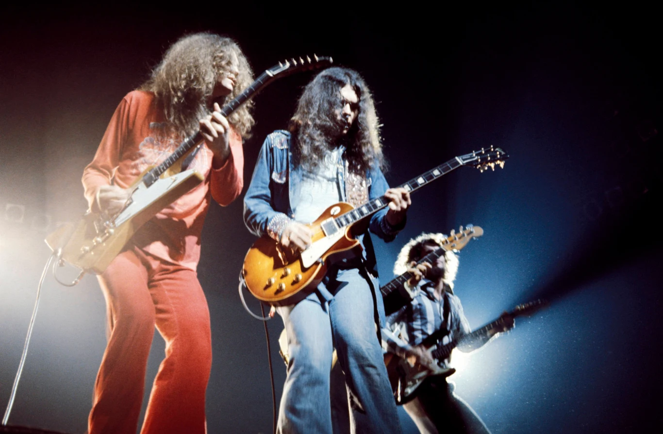 LYNYRD SKYNYRD Trzech gitarzystów grających na scenie podczas koncertu, intensywne oświetlenie sceniczne, skupienie na instrumentalistach i ich energii podczas występu.