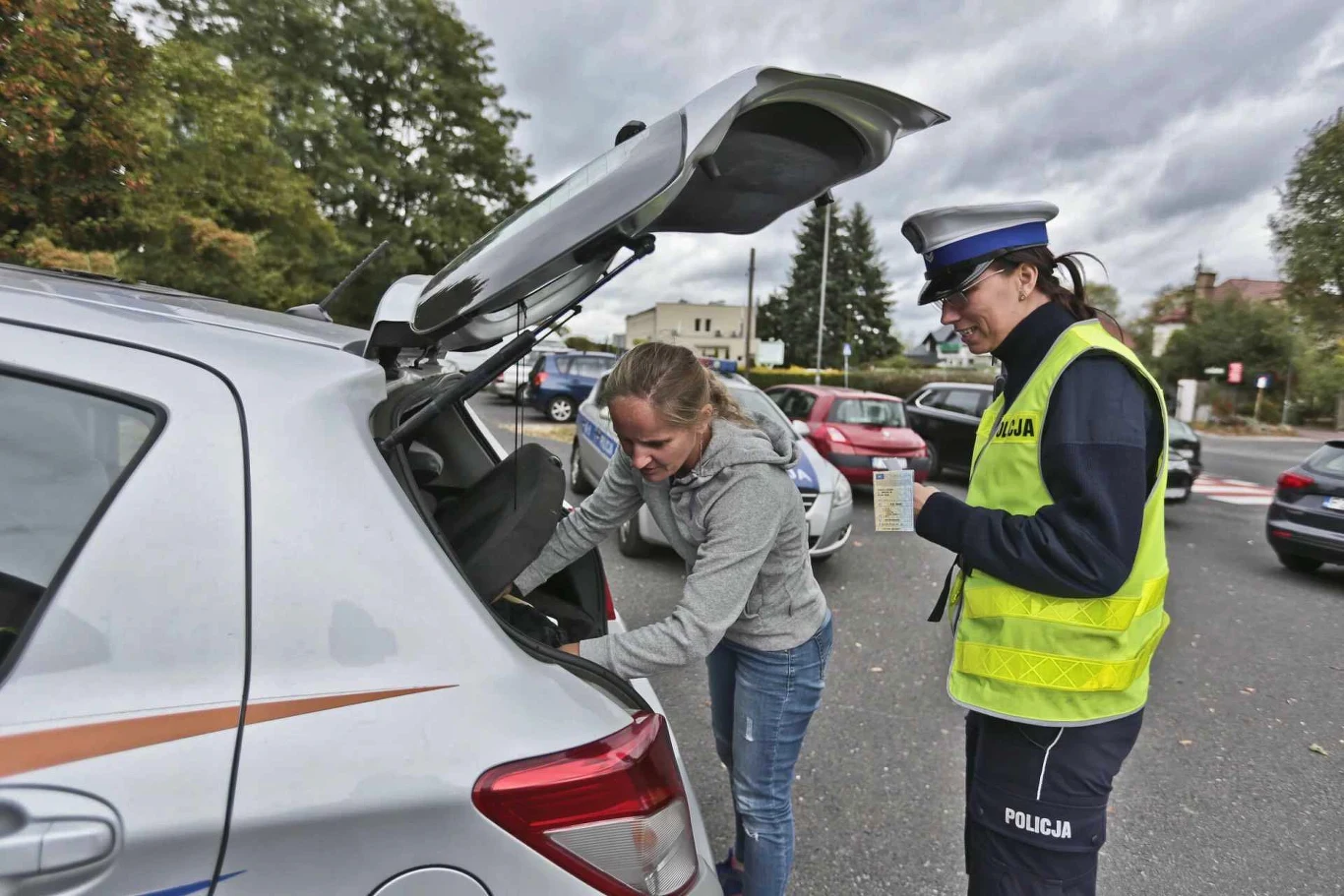 Kobieta otwiera bagażnik srebrnego samochodu na parkingu, obok stoi policjantka w odblaskowej kamizelce i mundurze, obserwując czynność, w tle zaparkowane auta i drzewa.