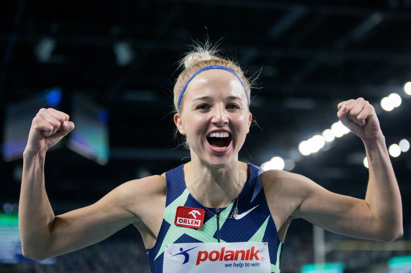 Karolina Kołeczek Zawodniczka ubrana w sportowy strój unosi obie zaciśnięte pięści w geście triumfu, szeroko się uśmiechając, w tle nienachalnie widoczna hala sportowa.
