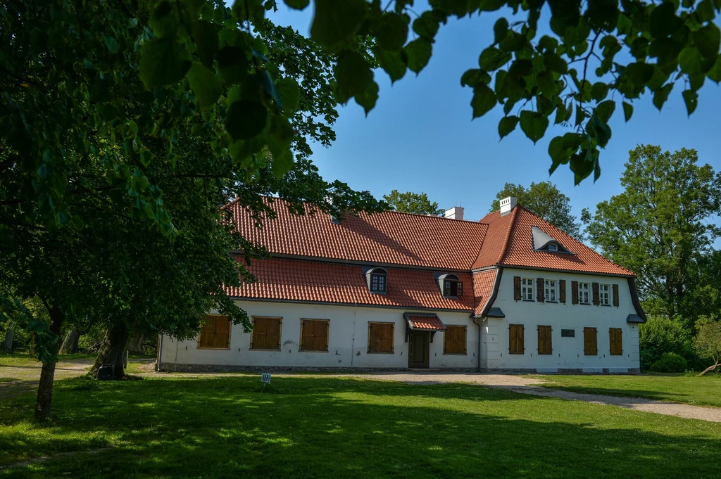 W Będominie urodził się Józef Wybicki - twórca słów "Mazurka Dąbrowskiego". Obecnie w dworze znajduje się interesujące muzeum Duży, zabytkowy dworek z czerwonym dachem, białą fasadą i zamkniętymi okiennicami, otoczony rozległym trawnikiem oraz drzewami, podkreślonym cieniem gałęzi na pierwszym planie.