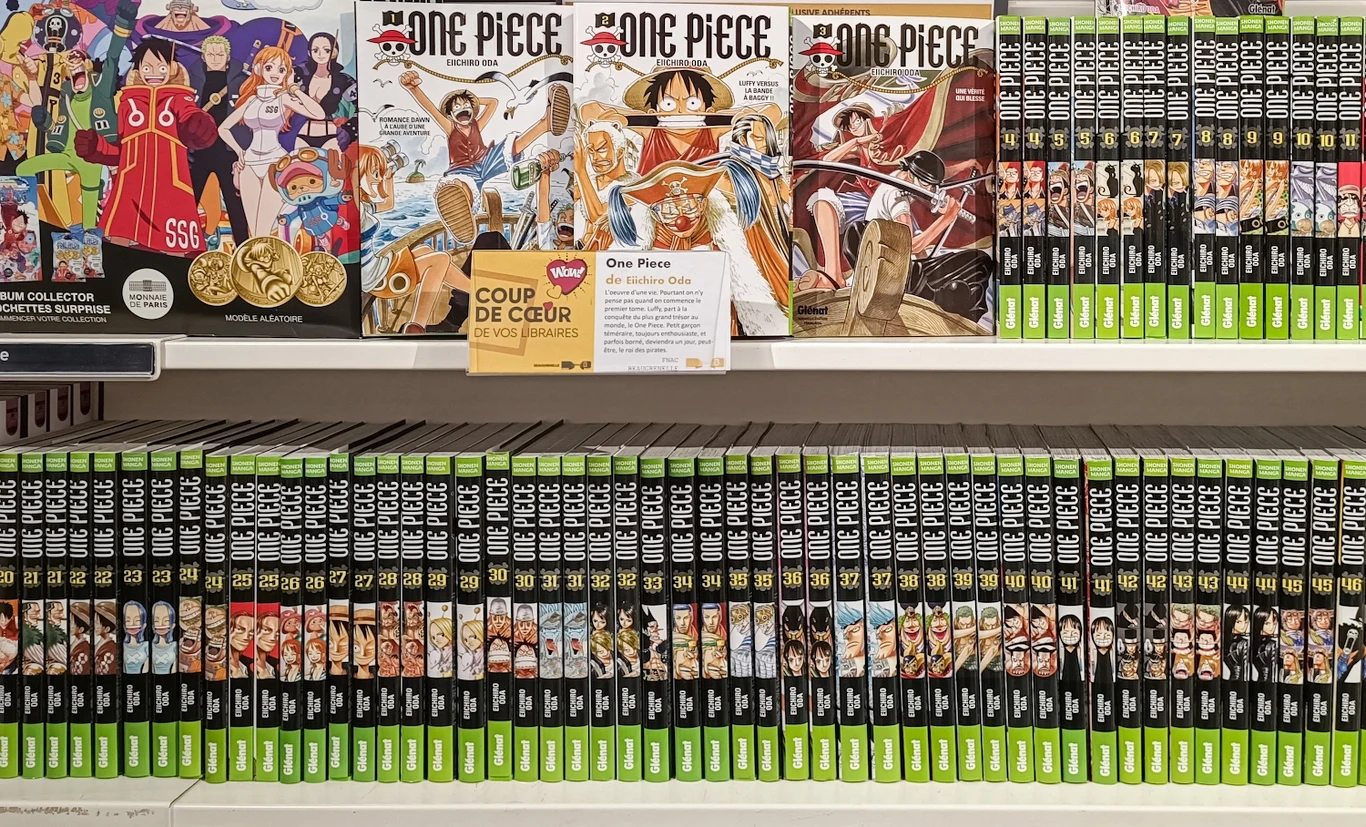 One Piece Kolekcja tomów mangi One Piece ułożonych na półkach, okładki z kolorowymi ilustracjami głównych bohaterów i materiałami promocyjnymi, na górnej półce widoczne są także dodatkowe gadżety związane z serią.