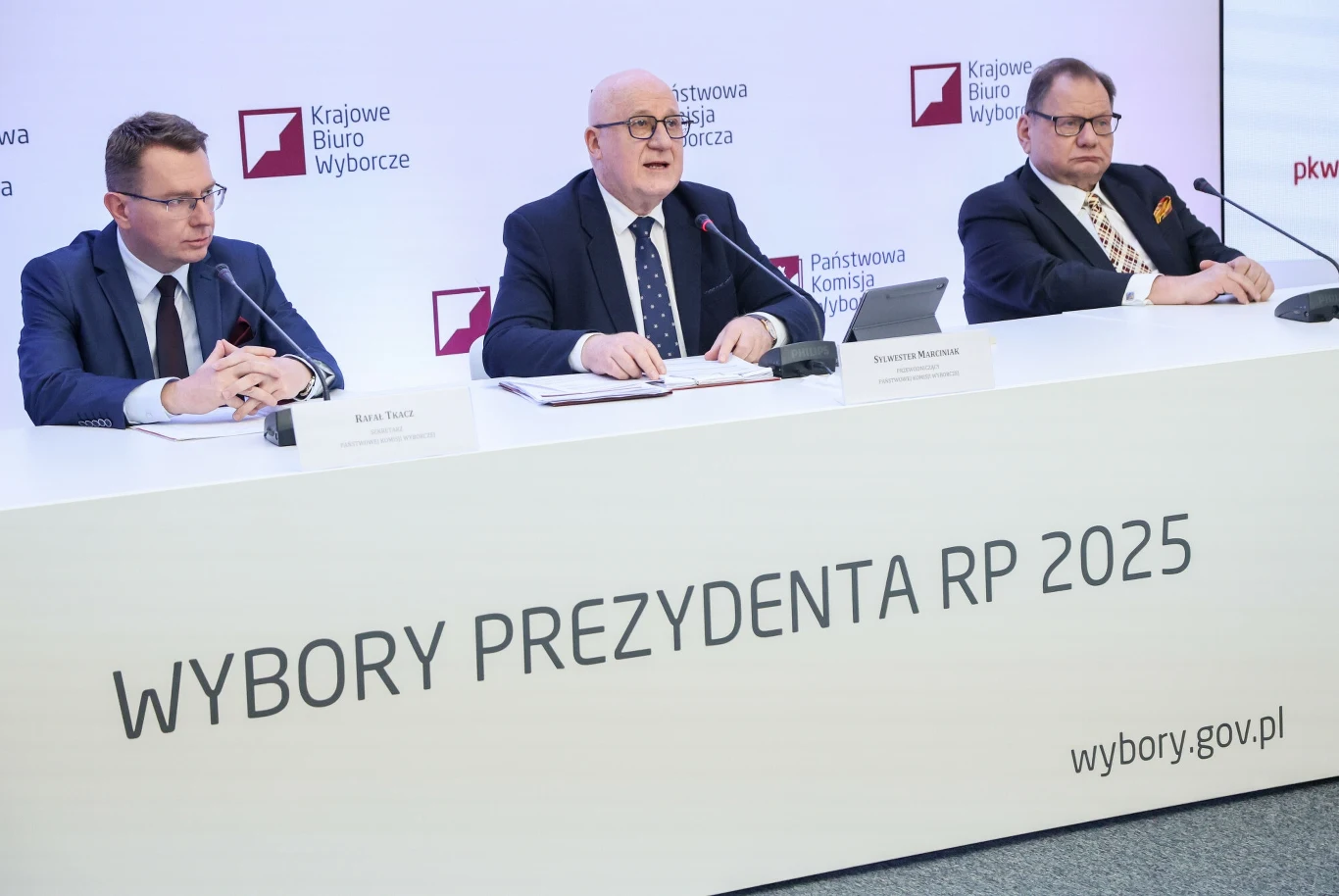 PKW podejmie działania w związku z zapowiedziami nieprawidłowości w niektórych z OKW Trzech mężczyzn w garniturach siedzi za stołem konferencyjnym podczas oficjalnej konferencji prasowej, na stole widoczne są mikrofony oraz tabliczki z nazwiskami, w tle logotypy Krajowego Biura Wyborczego oraz napisy związane z wyborami prezydenckimi 2...