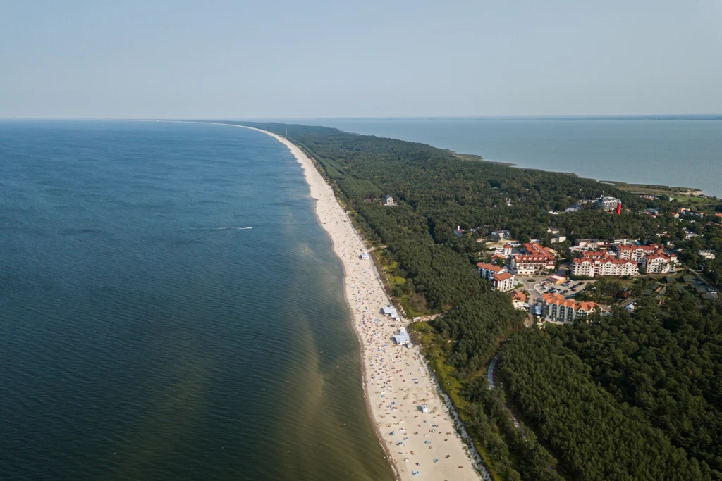 Krynica Morska to urokliwy kurort położony na Mierzei Wiślanej Szeroka, piaszczysta plaża ciągnąca się wzdłuż brzegu morza, po lewej stronie rozległa tafla wody, po prawej zwarte kompleksy leśne oraz zabudowania i hotele letniskowe, liczne osoby wypoczywające na plaży.