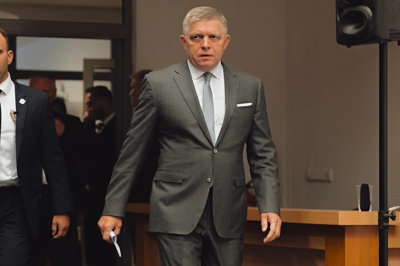 Premier Słowacji Robert Fico Mężczyzna w eleganckim garniturze wchodzi do pomieszczenia, trzymając dokument w ręku, za nim widocznych jest kilku innych ludzi, wnętrze urzędu lub sali konferencyjnej.