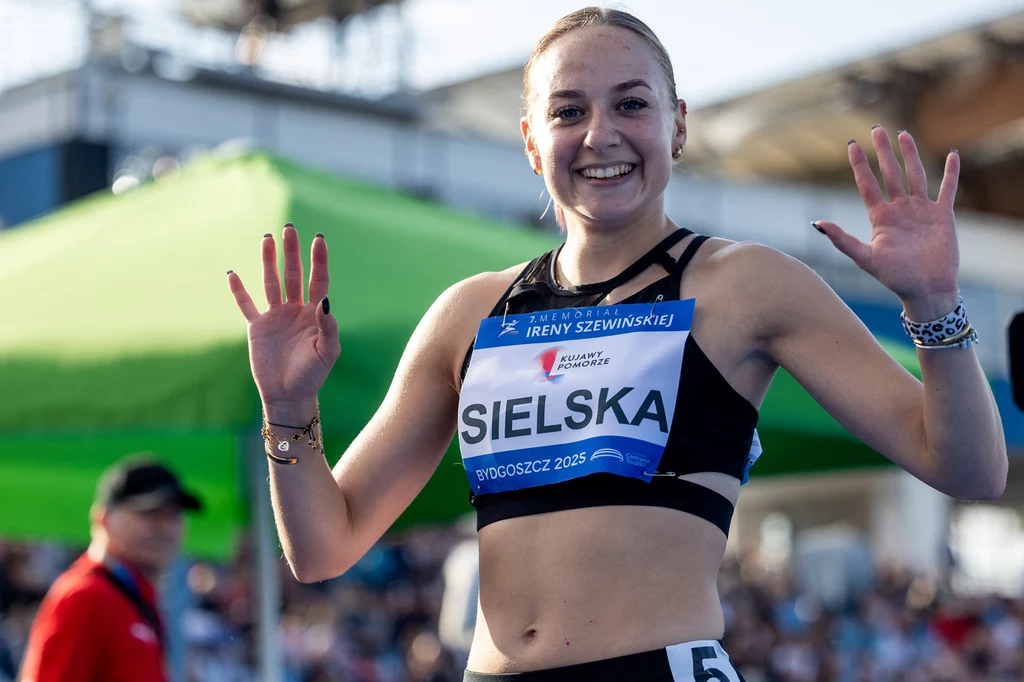 Młoda lekkoatletka w sportowym stroju uśmiecha się i unosi obie ręce, stojąc na stadionie podczas zawodów sportowych, wokół widoczne są elementy infrastruktury sportowej oraz inne osoby w tle.