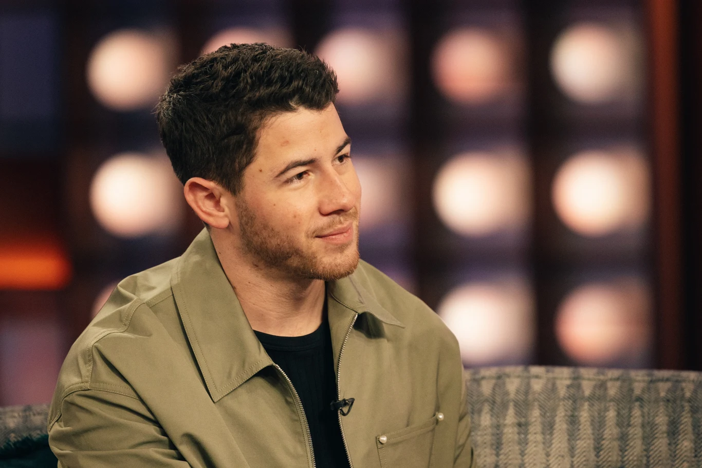 Nick Jonas ma zagrać Paula Stanleya w biografii zespołu Kiss Mężczyzna o krótkich, ciemnych włosach siedzi na kanapie w studiu telewizyjnym, ubrany w jasnobrązową kurtkę, w tle rozmyte światła sceniczne.