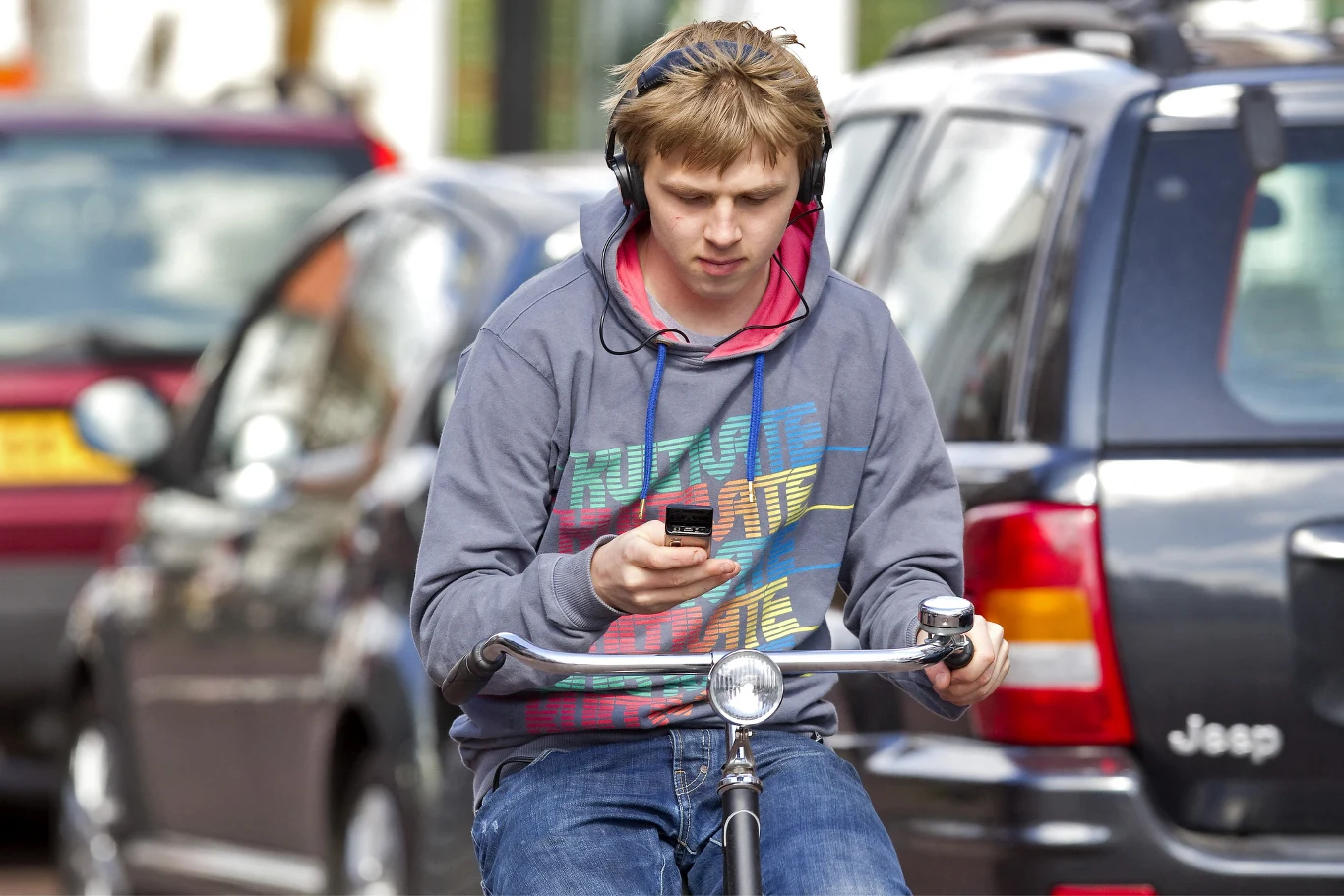 Przepisy stanowią jasno - podczas jazdy rowerem można słuchać muzyki przy wykorzystaniu słuchawek lub głośnika. Cykliści nie są jednak bezkarni. Młody mężczyzna jedzie rowerem ulicą między samochodami, ma na głowie słuchawki i patrzy w telefon trzymany w dłoni.